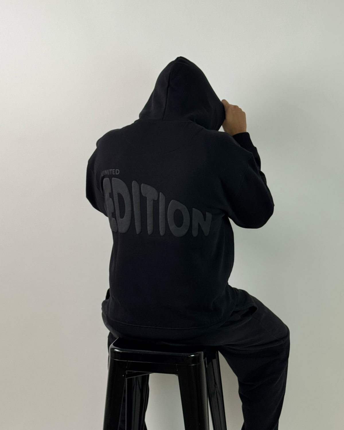 A person wearing a black hoodie and black pants, viewed from above while sitting on a black stool against a plain light gray background. The back of the hoodie has a large gray vertical text graphic reading 'CONDITION'.
