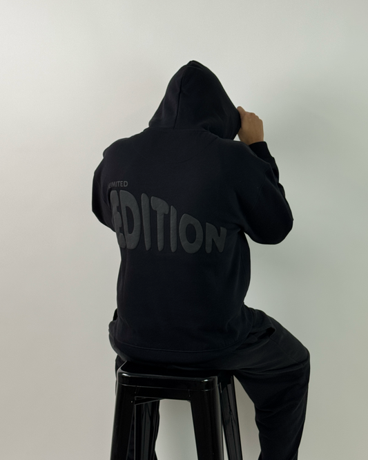 A person wearing a black hoodie and black pants, viewed from above while sitting on a black stool against a plain light gray background. The back of the hoodie has a large gray vertical text graphic reading 'CONDITION'.