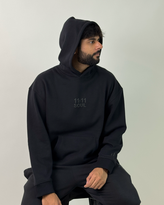 A man wearing a black hoodie and matching black sweatpants, shown from a side profile against a plain light gray background.