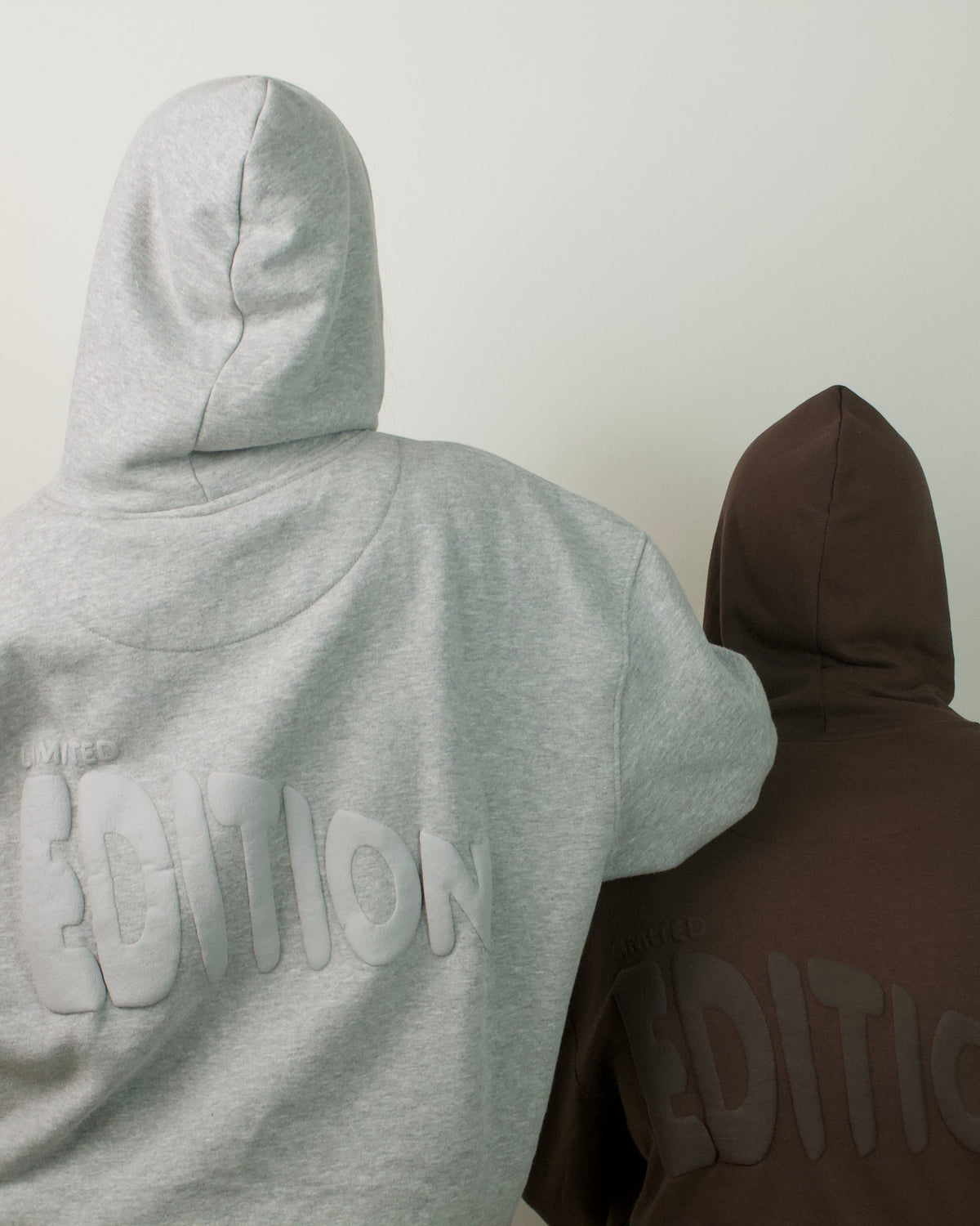 Two people seen from behind wearing hoodies, one light gray and one dark brown, standing against a plain light gray wall. The back of both hoodies features a large tonal 'EDITION' logo.