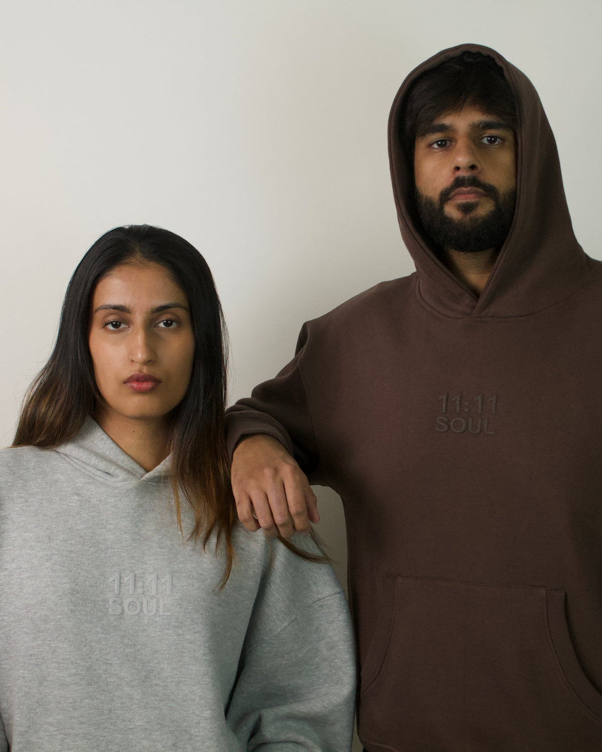A man and a woman standing side by side wearing matching oversized hoodies. The man wears a dark brown hoodie with a subtle tonal logo on the chest, and the woman wears a light gray hoodie with a matching tonal logo.