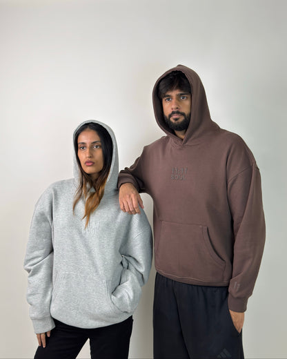 A man and a woman modeling oversized hoodies against a plain white wall. The woman wears a light gray heathered hoodie, and the man wears a brown hoodie with a tonal embroidered chest logo.
