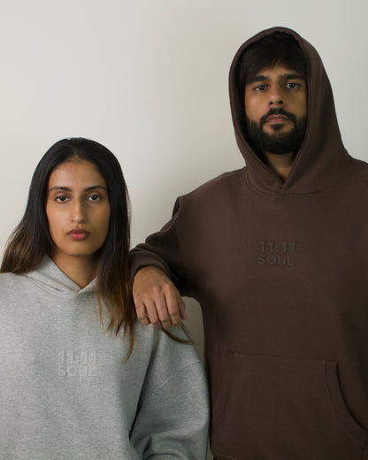 A man and a woman standing side by side wearing matching oversized hoodies. The man wears a dark brown hoodie with a subtle tonal logo on the chest, and the woman wears a light gray hoodie with a matching tonal logo.