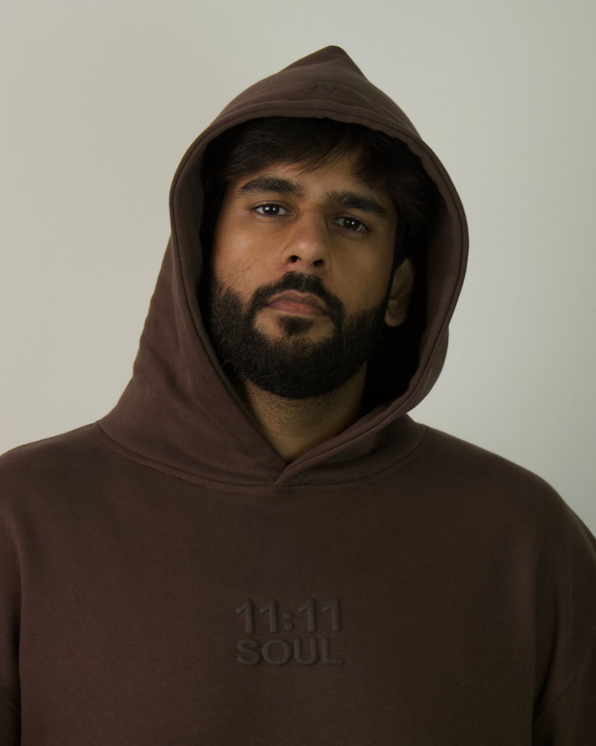 A man with a beard and dark hair wearing a dark brown pullover hoodie with '11.11 SOUL' printed on the chest, photographed against a plain light gray background.
