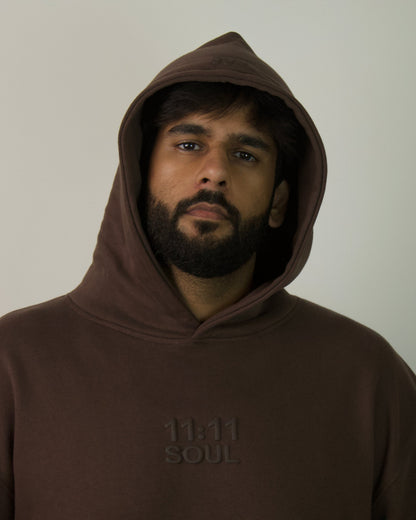 A man with a beard and dark hair wearing a dark brown pullover hoodie with '11.11 SOUL' printed on the chest, photographed against a plain light gray background.