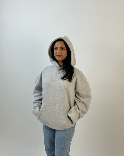 A woman wearing a light gray oversized pullover hoodie and blue jeans, photographed against a plain light gray wall.