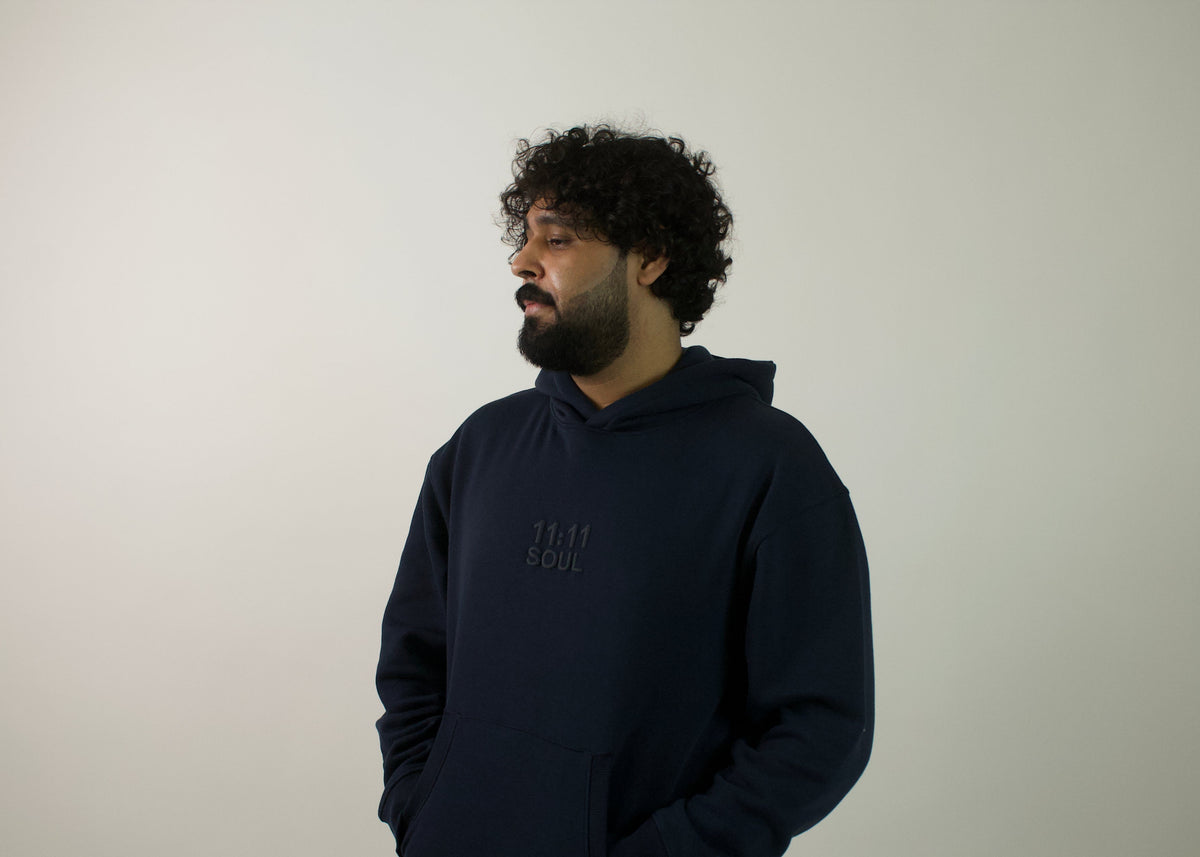 A man with curly dark hair and a beard wearing a dark navy blue hoodie with a tonal embroidered logo on the chest, standing against a plain light gray wall.