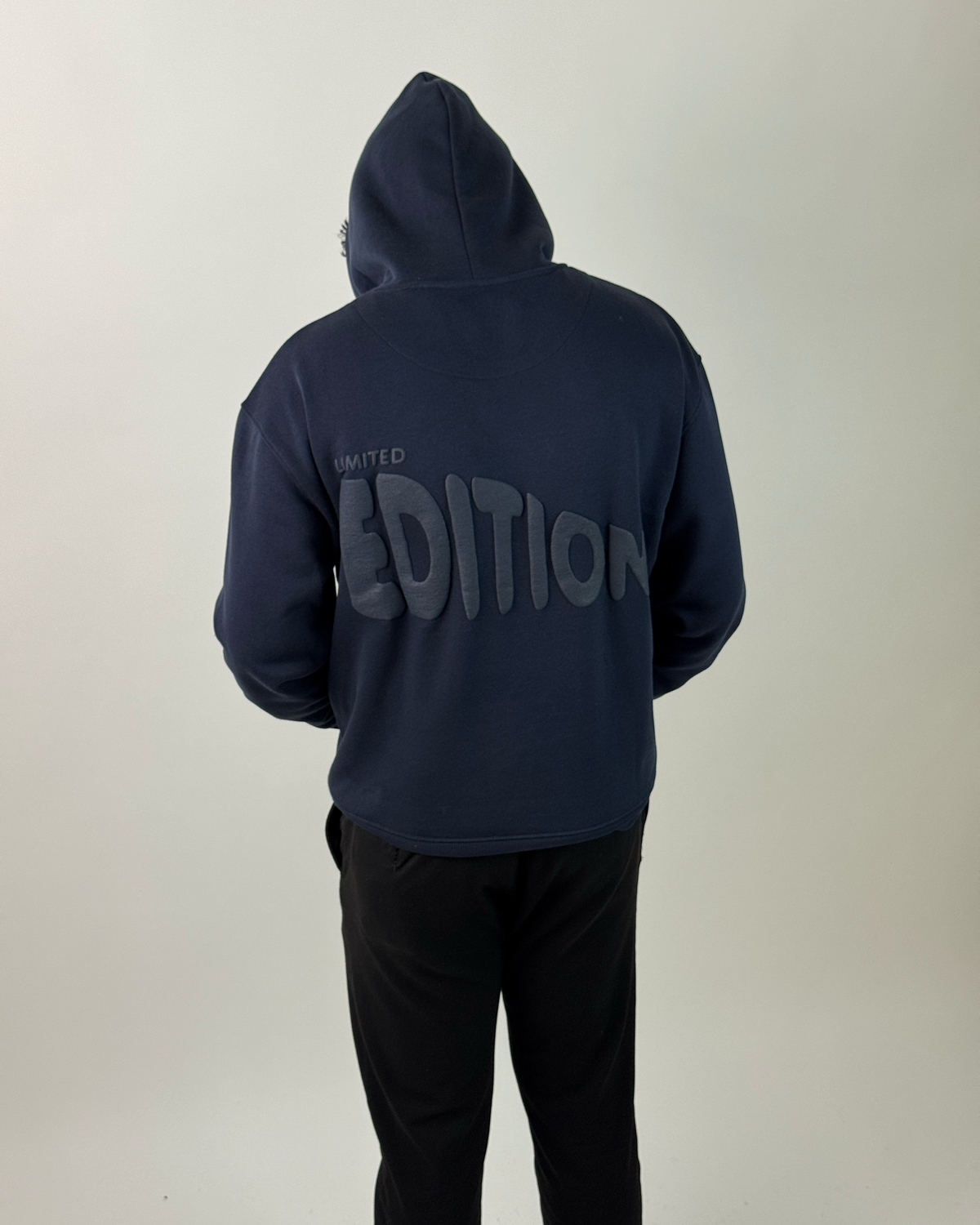 A person wearing a navy blue hoodie with a large dark gray 'EDITION' graphic on the back, paired with black pants, photographed against a plain light gray background.