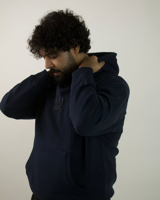 A man with curly dark hair and a beard wearing a navy blue hoodie, adjusting the collar with one hand while looking down. The hoodie has a small dark graphic on the left chest.