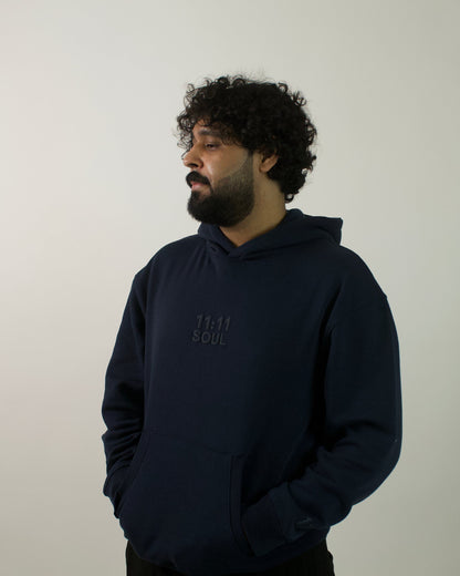 A man with curly dark hair and a beard wearing a dark navy blue hoodie with a tonal embroidered logo on the chest, standing against a plain light gray wall.