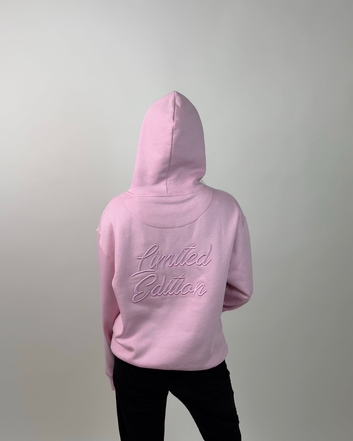 A person shown from the back wearing a light pink hoodie with 'Limited Edition' embroidered in matching pink script across the upper back, paired with black pants against a plain light gray studio background.