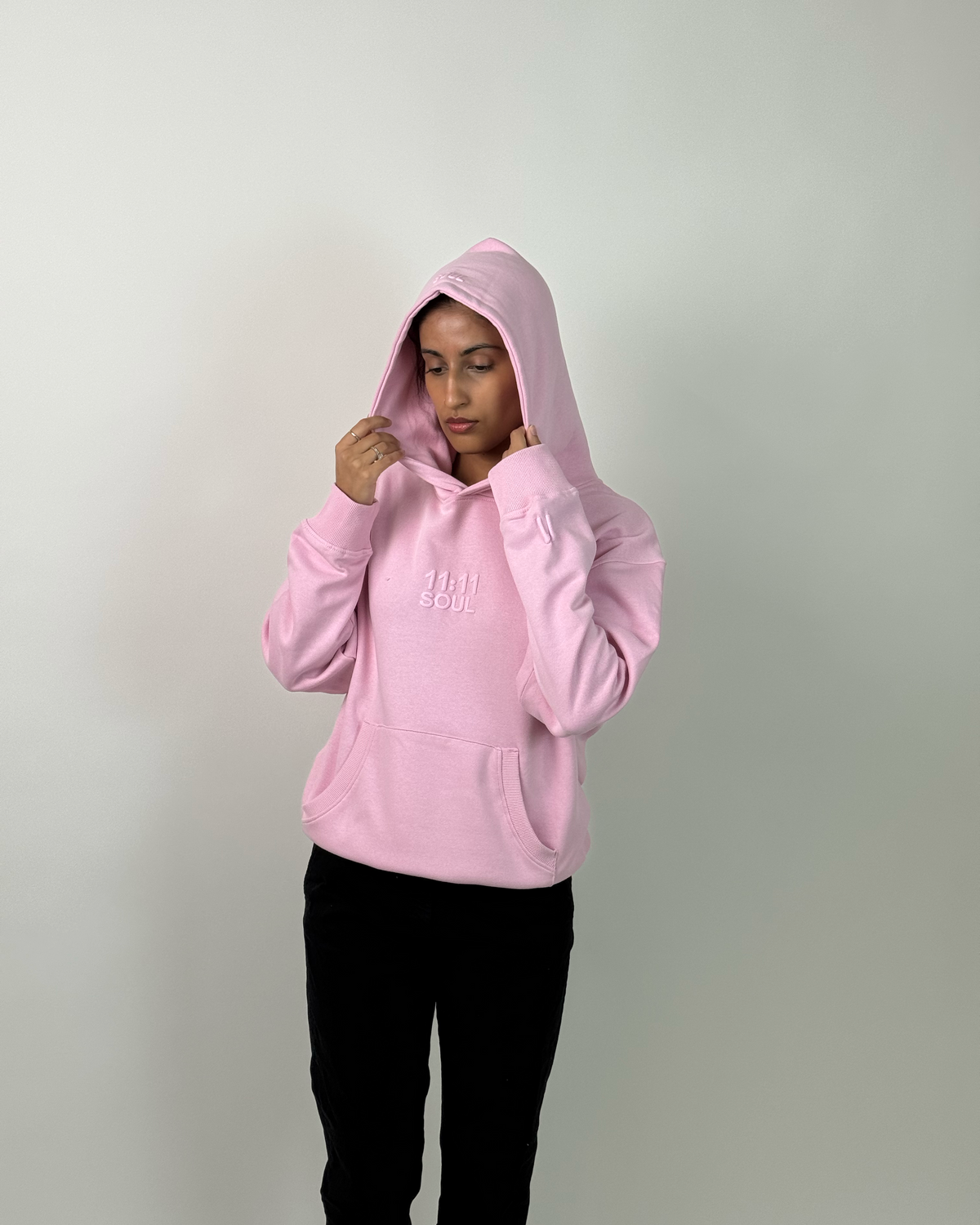 A person wearing a light pink pullover hoodie with a small white embroidered logo on the chest, paired with black pants, shown against a plain light gray background.