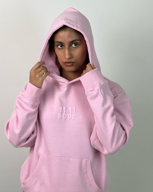 A person wearing a light pink pullover hoodie with a tonal embroidered logo on the chest, posed against a plain light gray background.